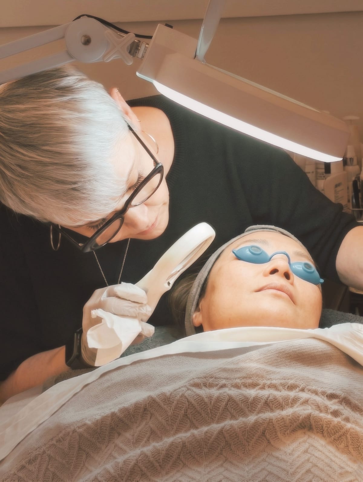 Jill Jodar performing a facial treatment under a magnifying lamp while the client wears protective eye shields