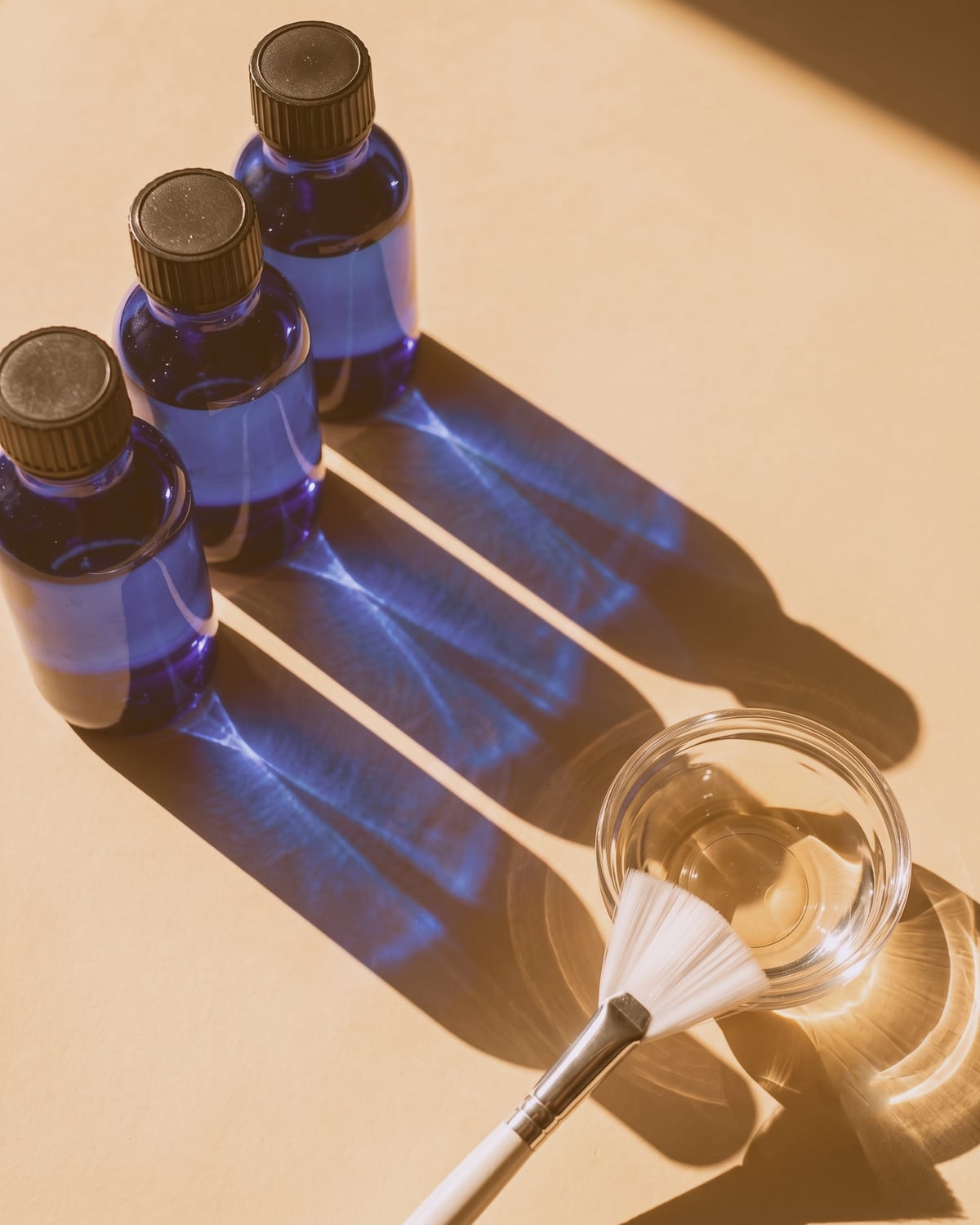 Three cobalt blue glass bottles casting long blue shadows in strong side light, beside a glass dish and fan brush on a warm surface